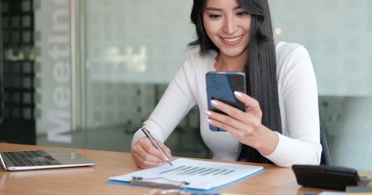 Woman looking at her phone while writing down notes on a paper